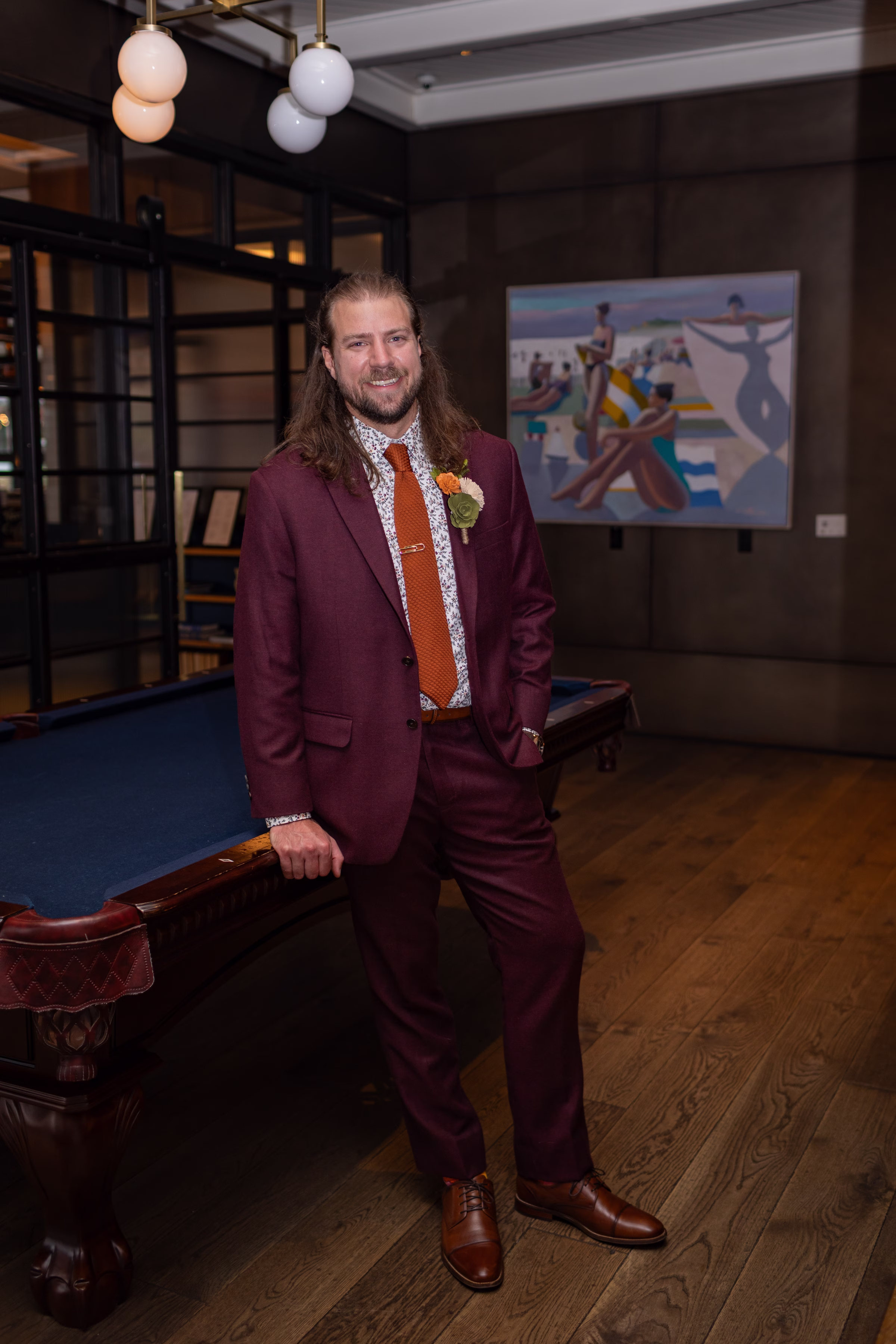 moody romantic fall wedding loft on broadway milwaukee wisconsin groomsmen 40 0110 - A Moody, Romantic Fall Wedding at The Loft on Broadway | Sarah + Nathan a groom poses, leaning against a pool table. He is wearing a burgundy suit from Sola Wood Flowers with a floral patterned dress shirt, burnt orange tie, brown shoes, and a wood flower boutonniere