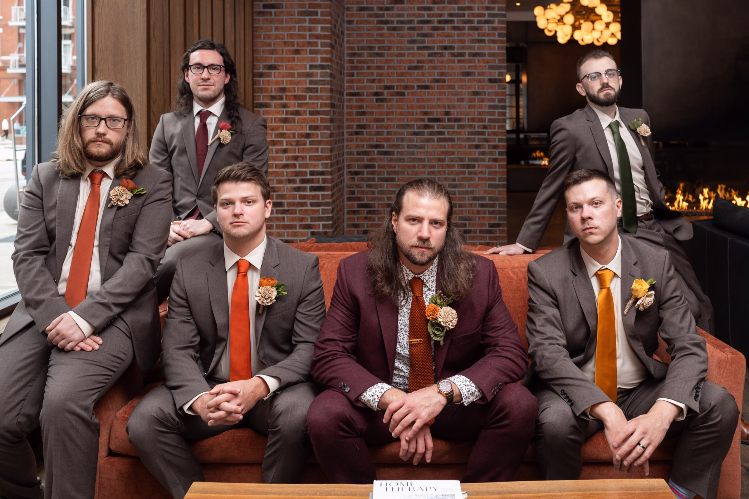 moody romantic fall wedding loft on broadway milwaukee wisconsin groomsmen 48 0135 - A Moody, Romantic Fall Wedding at The Loft on Broadway | Sarah + Nathan a groom sits, surrounded by his groomsmen for a serious portrait. He is wearing a burgundy suit from Studio Suits, with floral patterned dress shirt, and a burnt orange tie. Groomsmen are dressed in dark gray suits with white dress shirts, ties matching the bridesmaids dresses, and Sola Wood Flowers boutonnieres