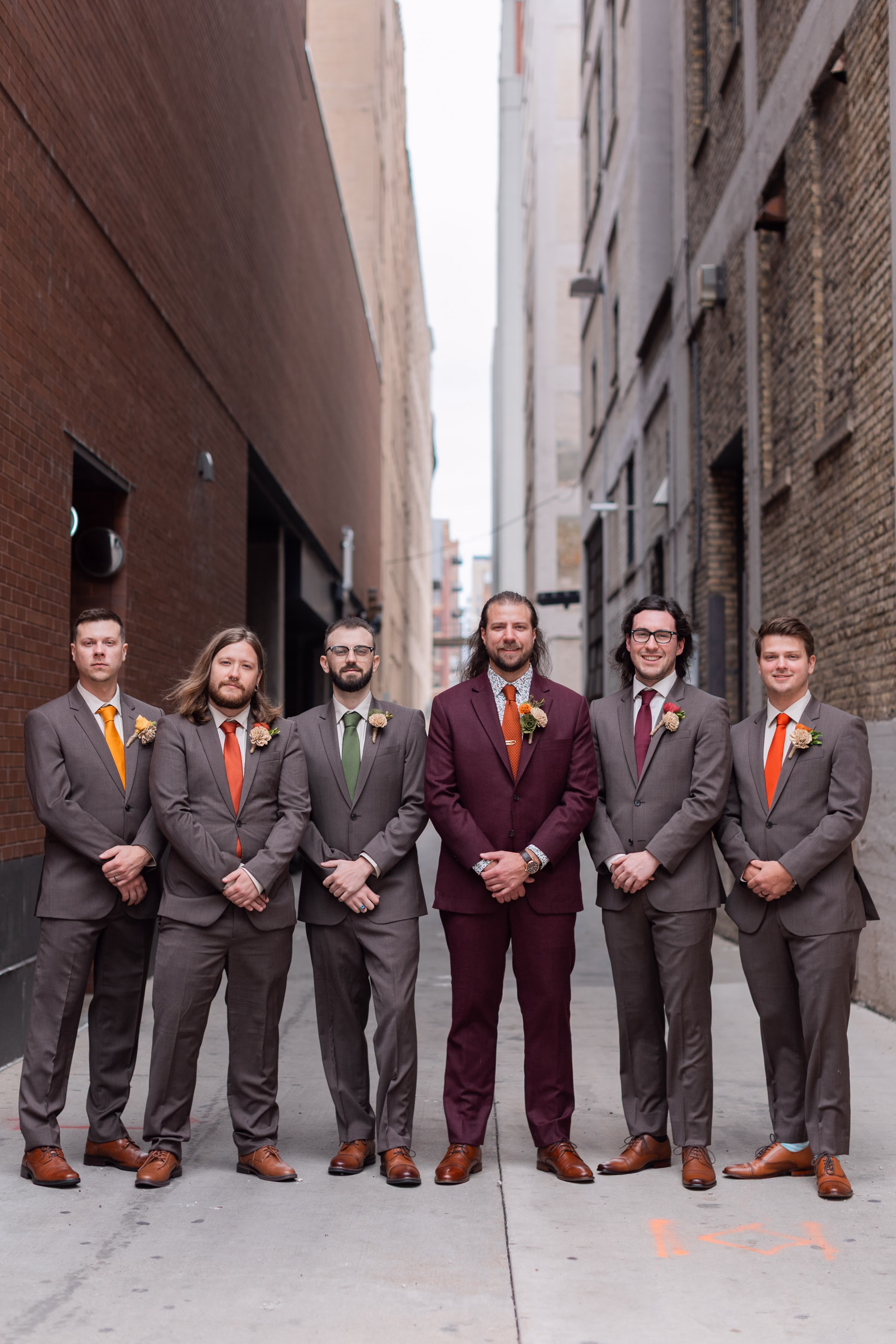 moody romantic fall wedding loft on broadway milwaukee wisconsin groomsmen 53 10105 - A Moody, Romantic Fall Wedding at The Loft on Broadway | Sarah + Nathan a groom and his groomsman stand side-by-side in an alley with brick walls on either side in Milwaukee, Wisconsin. They are wearing suits from Studio Suits, with the groom in burgundy and his groomsmen in dark gray