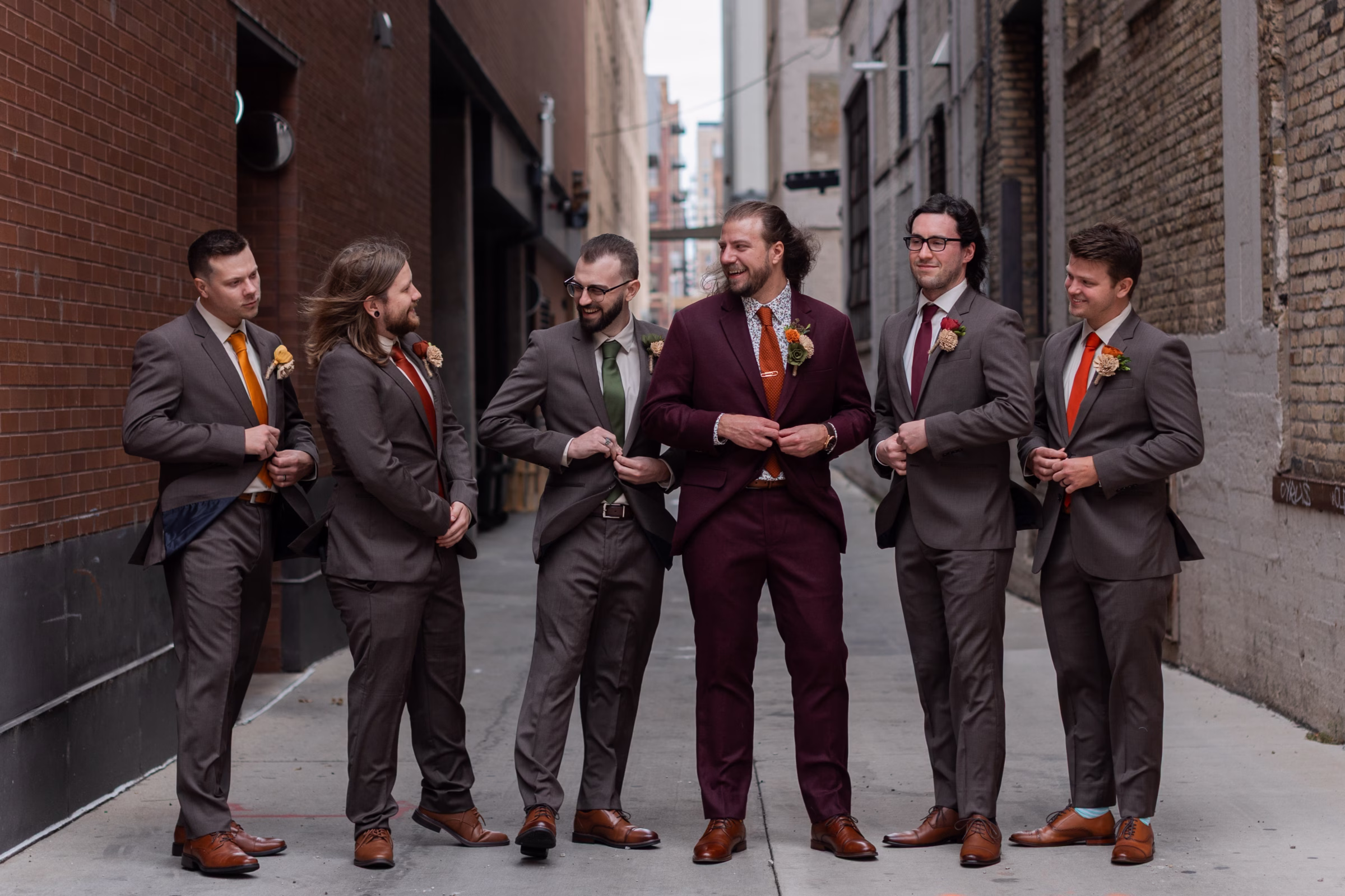 moody romantic fall wedding loft on broadway milwaukee wisconsin groomsmen 60 10147 - A Moody, Romantic Fall Wedding at The Loft on Broadway | Sarah + Nathan a groom and his groomsmen stand in an alley surrounded by brick buildings and Milwaukee, Wisconsin architecture. They are casually interacting and buttoning their suit jackets from Studio Suits