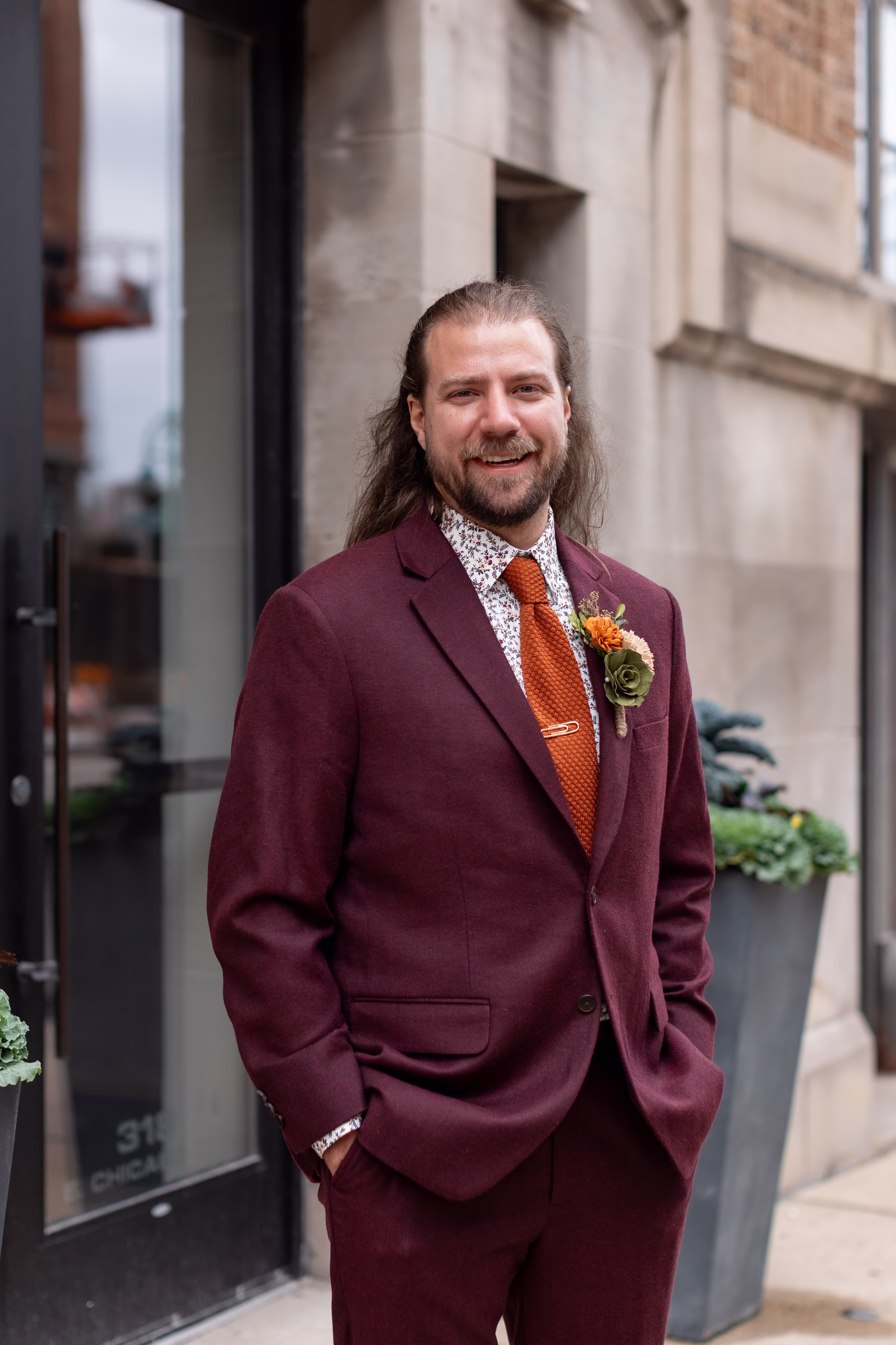 moody romantic fall wedding loft on broadway milwaukee wisconsin groomsmen 71 10179 - A Moody, Romantic Fall Wedding at The Loft on Broadway | Sarah + Nathan a groom stands with his hands in the pockets of his burgundy suit from Studio Suits. He is also wearing a patterned dress shirt, a burnt orange tie, and a wood flower boutonnière