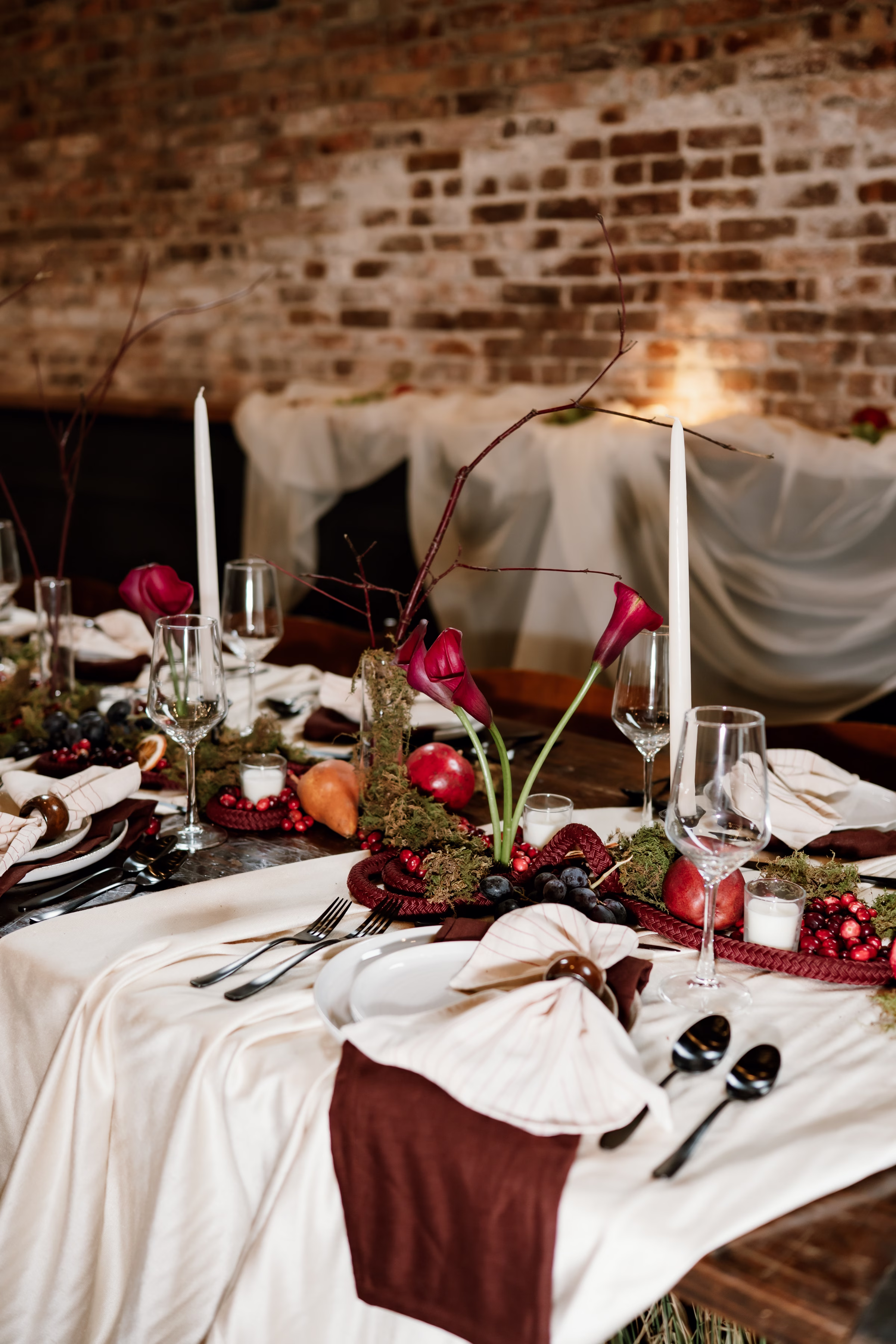 CorrinaNicolePhotography AmandaJenPhotography MercantileHall StyledShoot 112825 4 - Rich Velvet Fall Wedding Inspiration at Mercantile Hall in Burlington, Wisconsin a close-up of a wedding reception tablescape showing a luxurious white tablecloth layered with white plating, crystal glassware, candles, fall fruit as décor, burgundy florals, and mossy greenery