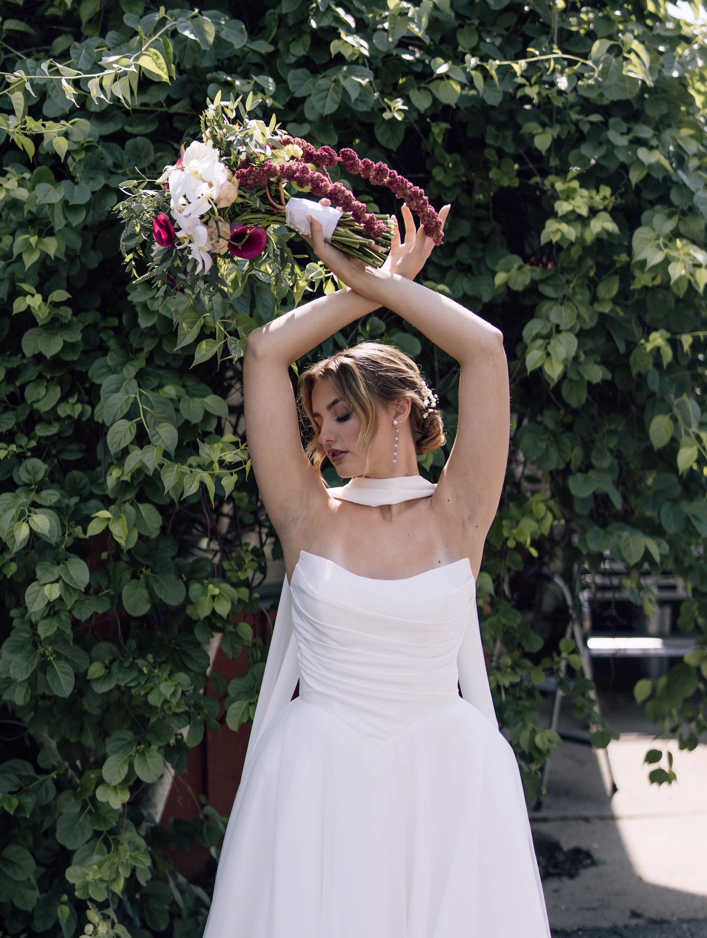A Wisconsin bride posing elegantly in front of a lush greenery wall at a wedding planned by Northwoods Event Services, holding a dramatic bouquet of cascading purple blooms, white orchids, and deep pink calla lilies.