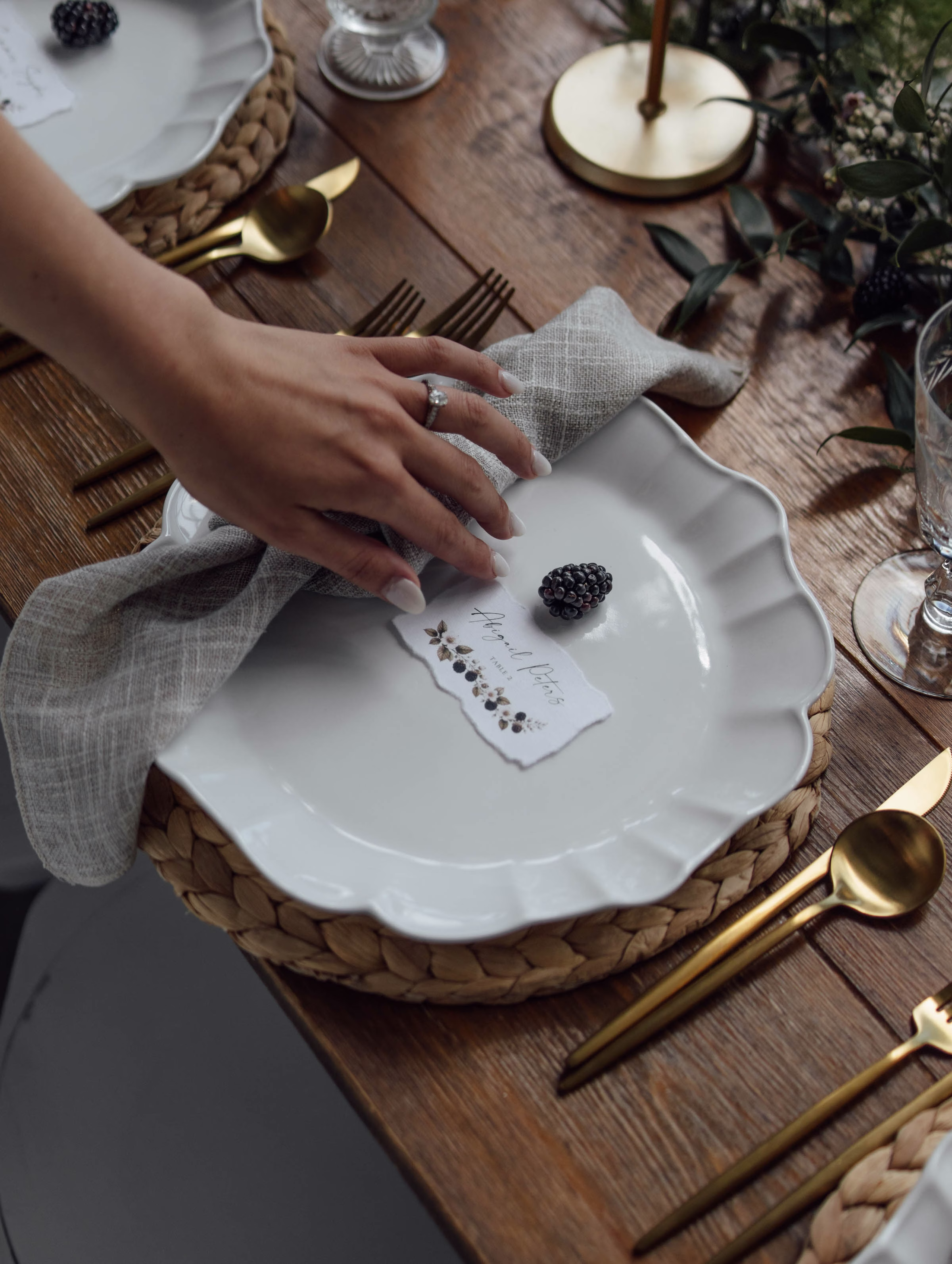A rustic-chic wedding place setting styled by Northwoods Event Services featuring a white scalloped plate on a woven charger, a textured gray linen napkin, matte gold flatware, and a custom name card topped with a fresh blackberry.