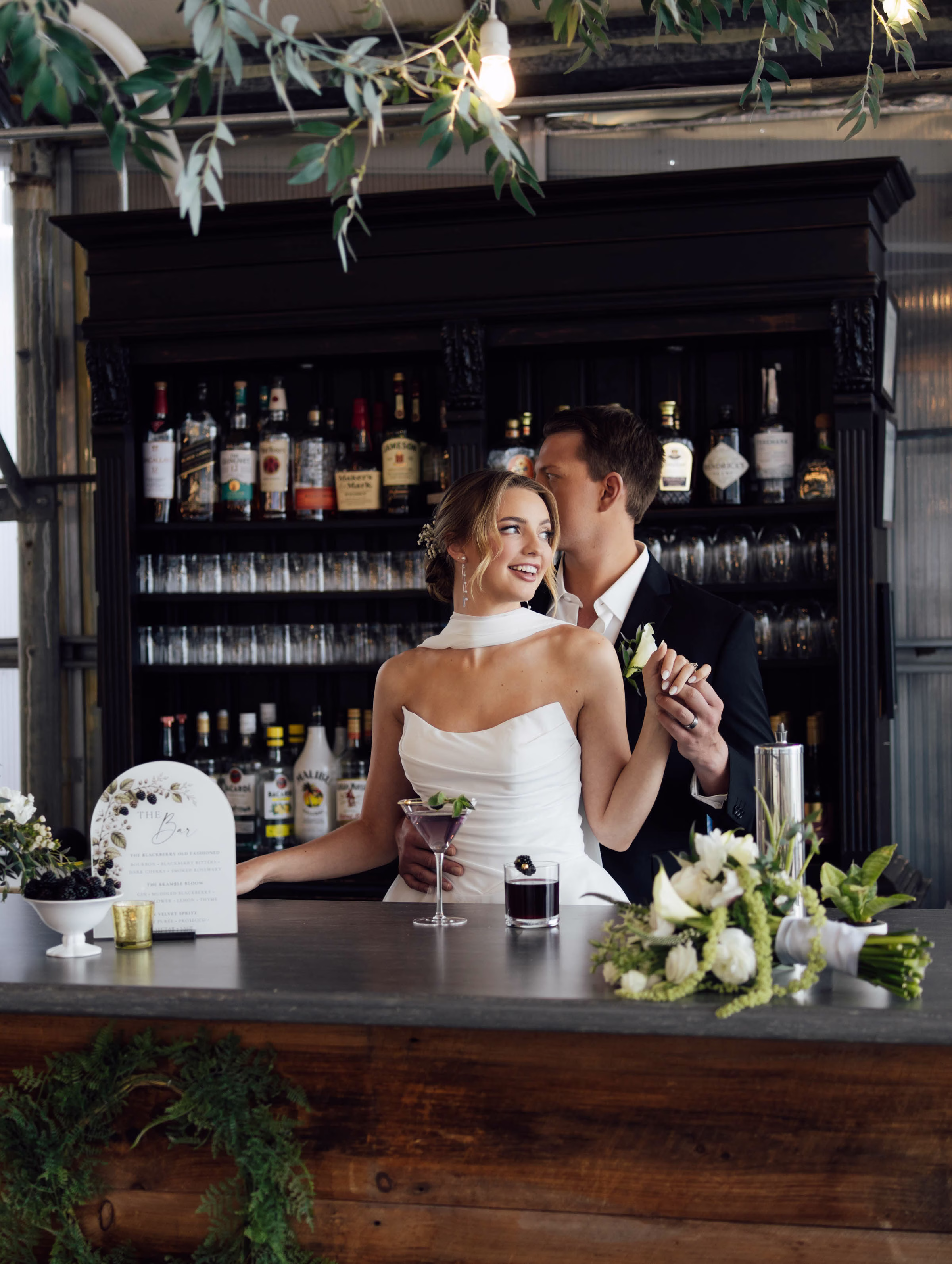 A customized wedding reception bar area planned by Northwoods Event Services, featuring a dark wood back bar stocked with liquor bottles and a couple smiling together behind a rustic wood bar counter with signature cocktails.