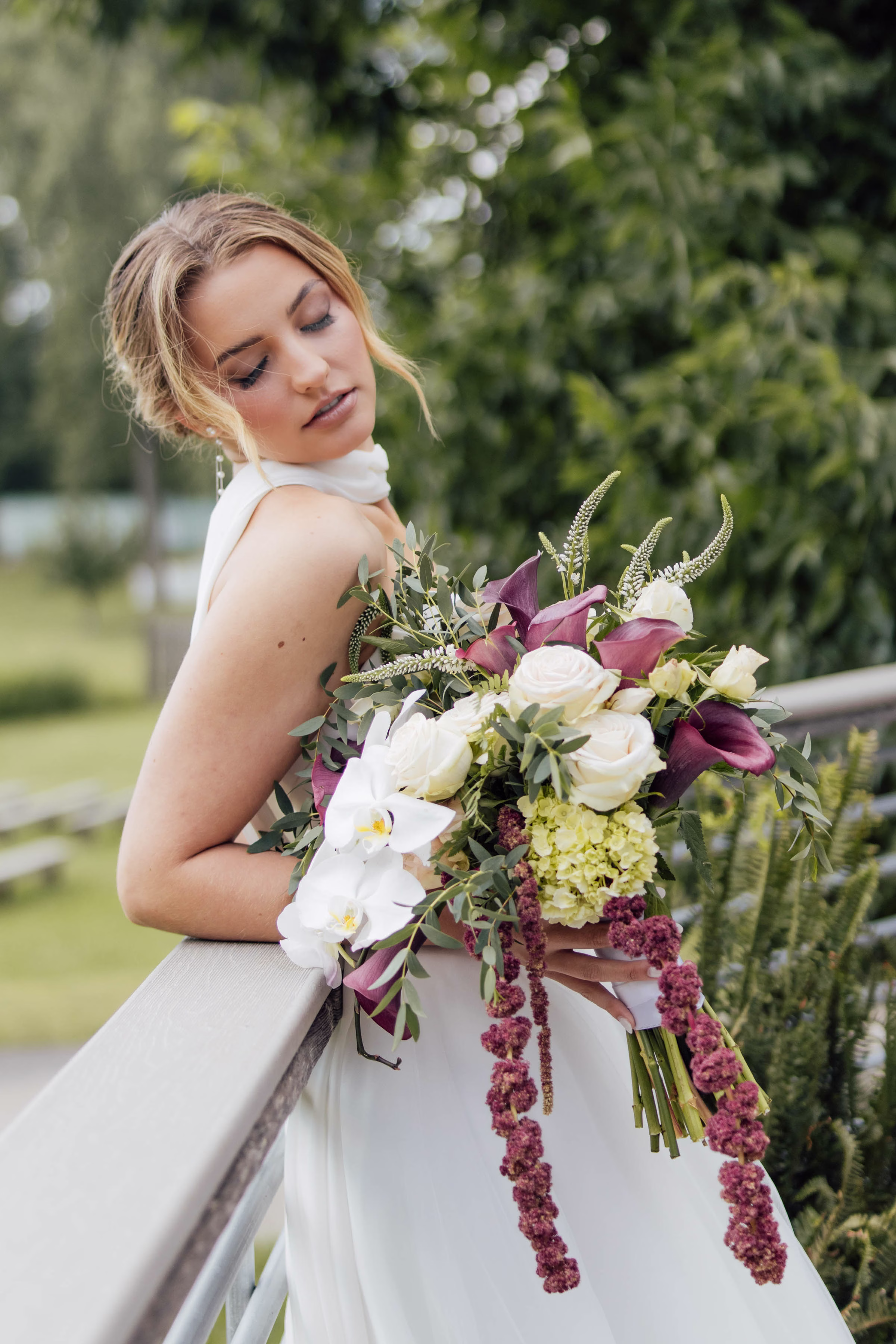 A Wisconsin bride leaning on a railing while showcasing a lush wedding bouquet coordinated by Northwoods Event Services, featuring cascading amaranthus, white orchids, white roses, and purple calla lilies.