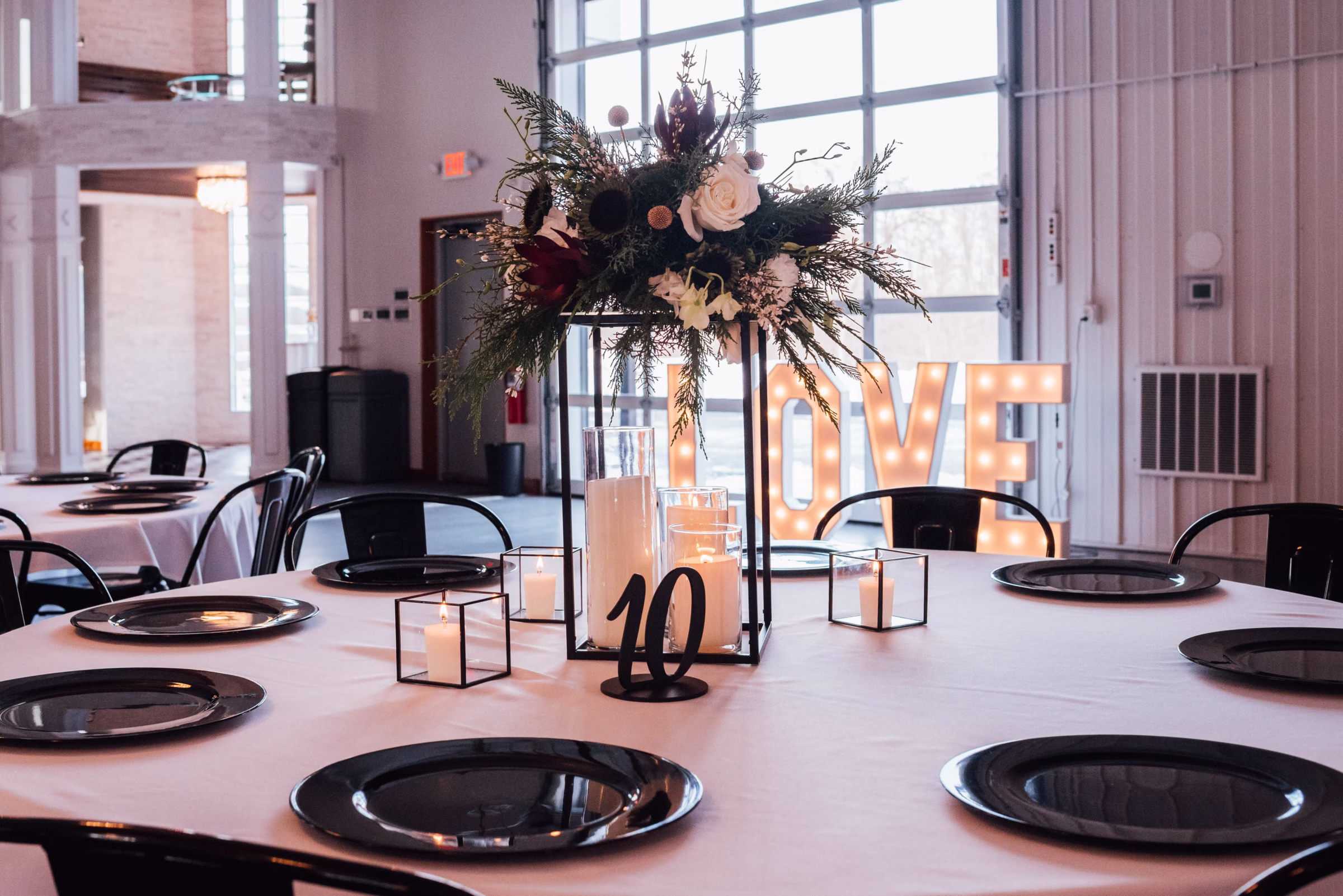 A modern industrial wedding reception table designed by Northwoods Event Services featuring black charger plates, glass candle cylinders, and a tall geometric metal centerpiece stand overflowing with winter greenery and white roses, with large marquee "LOVE" letters glowing in the background.