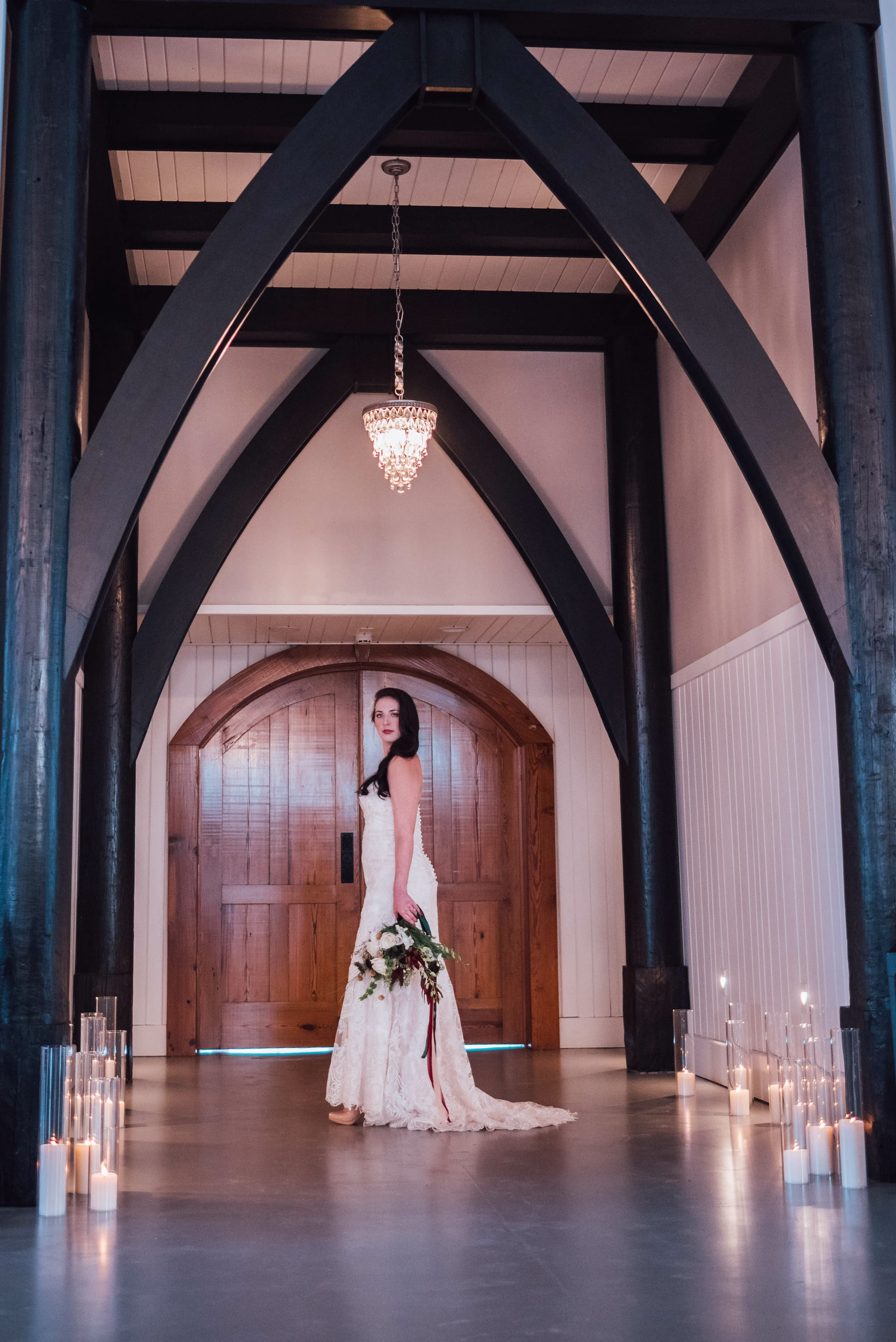 A striking bridal portrait from a Wisconsin wedding coordinated by Northwoods Event Services, showcasing a bride in a lace gown standing under dramatic black timber framing and a crystal chandelier, flanked by glowing glass cylinder candles.
