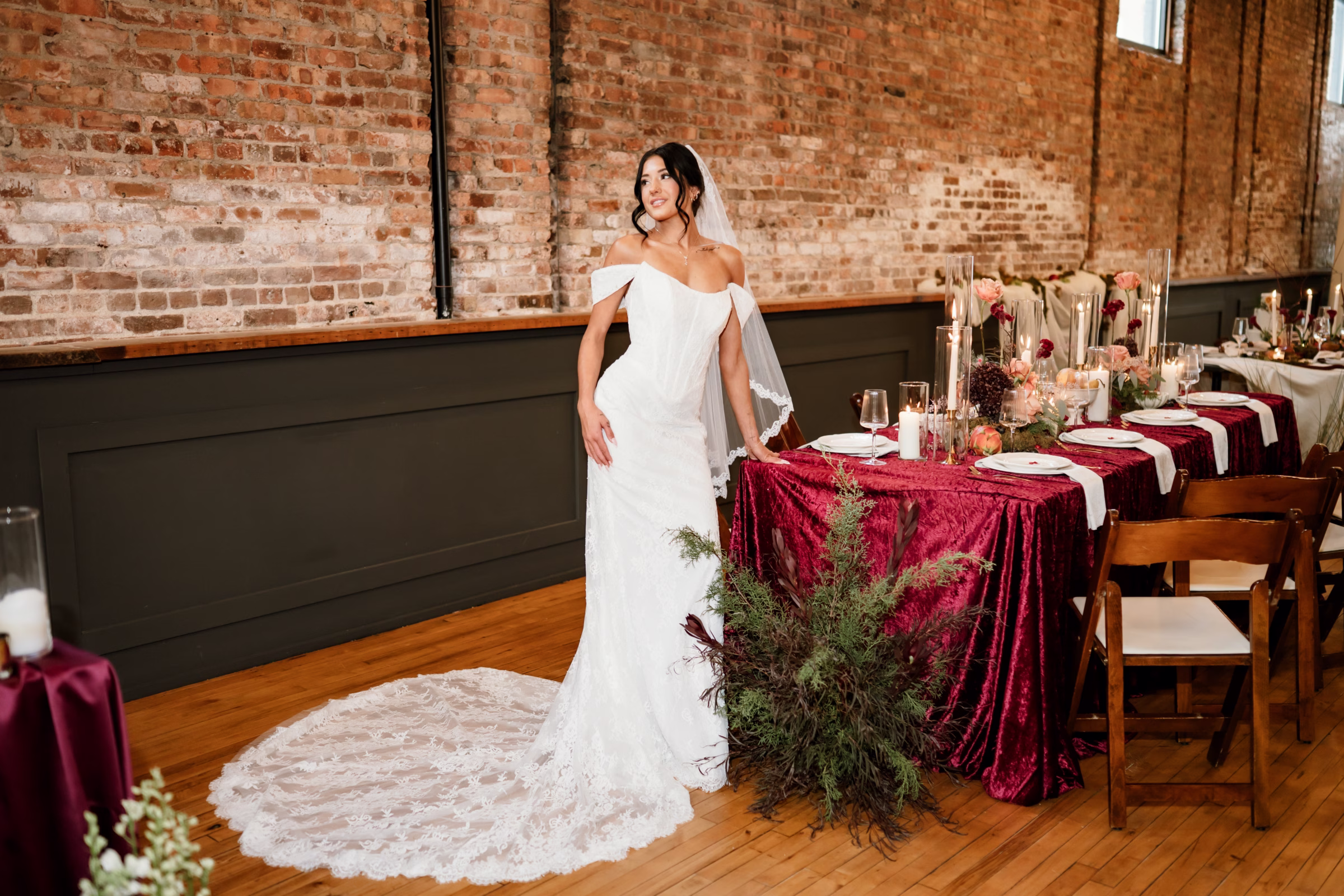 rich velvet fall wedding inspiration mercantile hall burlington wisconsin burgundy tablescape bride fashion 219 - Rich Velvet Fall Wedding Inspiration at Mercantile Hall in Burlington, Wisconsin a bride poses at the end of a reception table showing off her dress from Bon Bon Belle Bridal boutique, along with the natural brick walls of Mercantile Hall
