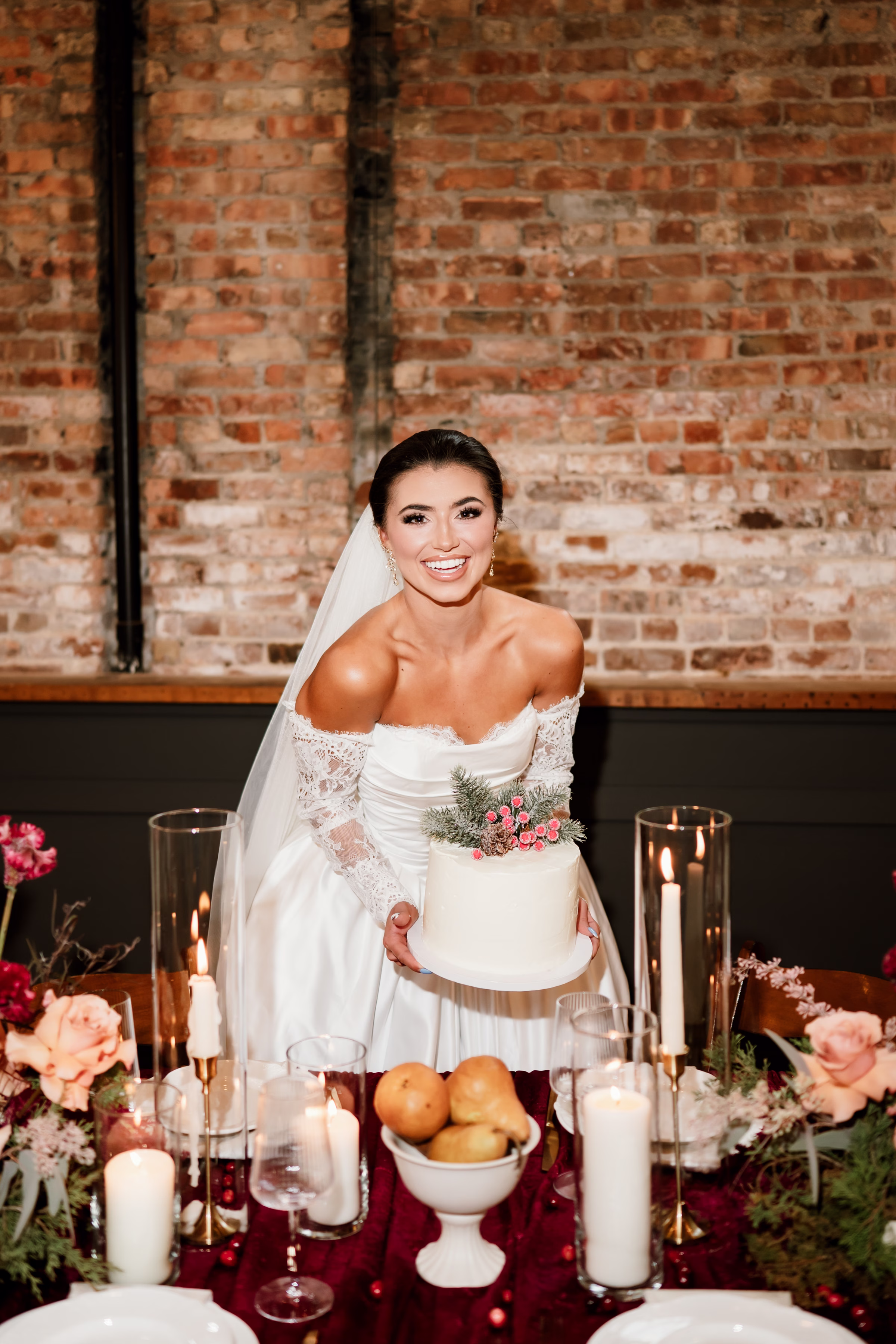 rich velvet fall wedding inspiration mercantile hall burlington wisconsin cake fruit tablescape 239 - Rich Velvet Fall Wedding Inspiration at Mercantile Hall in Burlington, Wisconsin a bride smiles while holding a single-layer wedding cake by Kindred Bakeshop topped with seasonal pine branches, a pine cone, and winter berries. Mercantile Hall's natural brick backdrop balances against a tablescape with rich burgundy velvet tablecloth