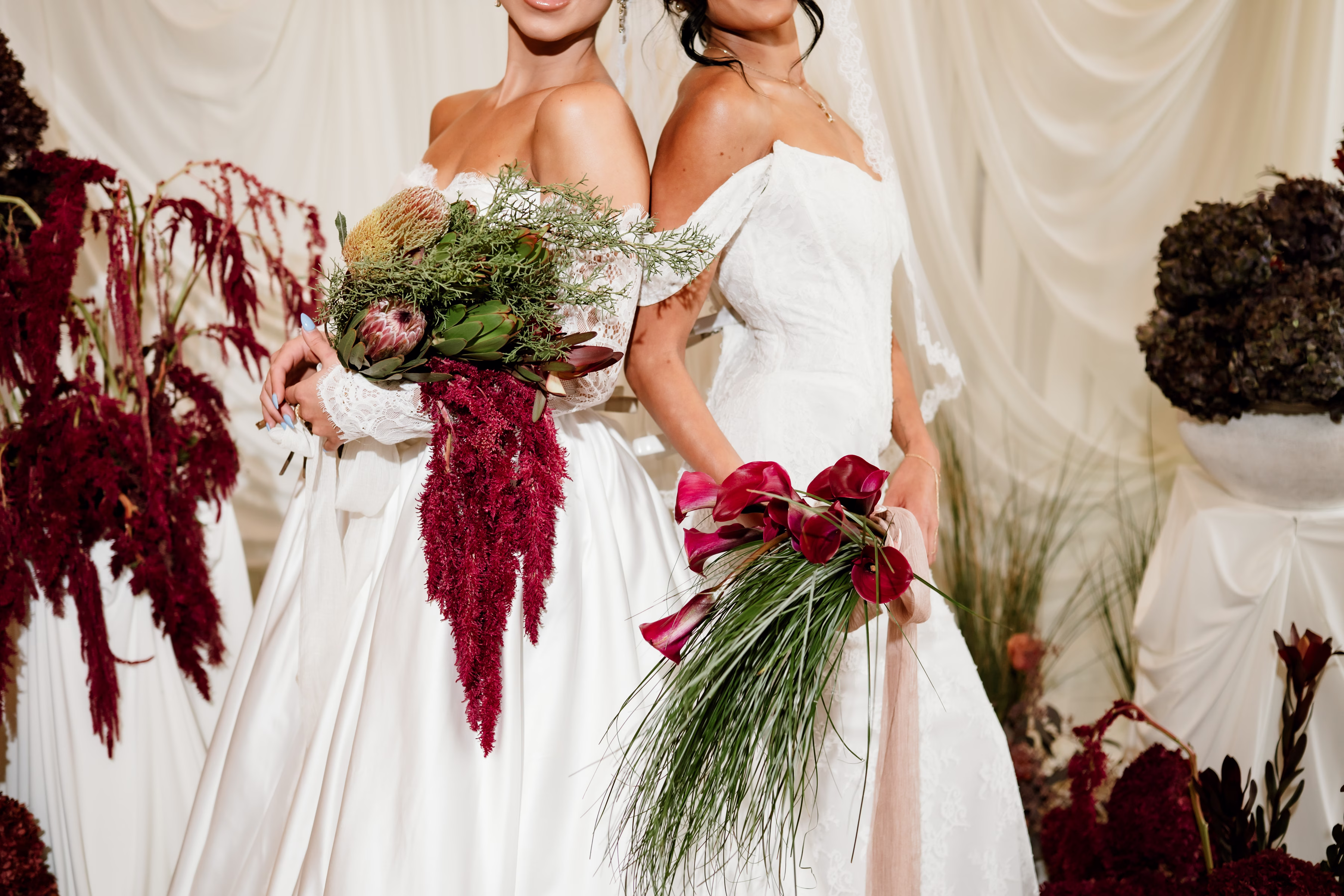 rich velvet fall wedding inspiration mercantile hall burlington wisconsin ceremony draping bride fashion 154 - Rich Velvet Fall Wedding Inspiration at Mercantile Hall in Burlington, Wisconsin two brides stand back-to-back, each holding fall seasonal bouquets from The Flower Bar and Anna's Flora. The left bouquet includes king protea, artichoke, leucadendron, and cascading burgundy amaranthus accented with delicate branching greenery. The right bouquet pairs deep burgundy calla lilies with cascading bear grass.