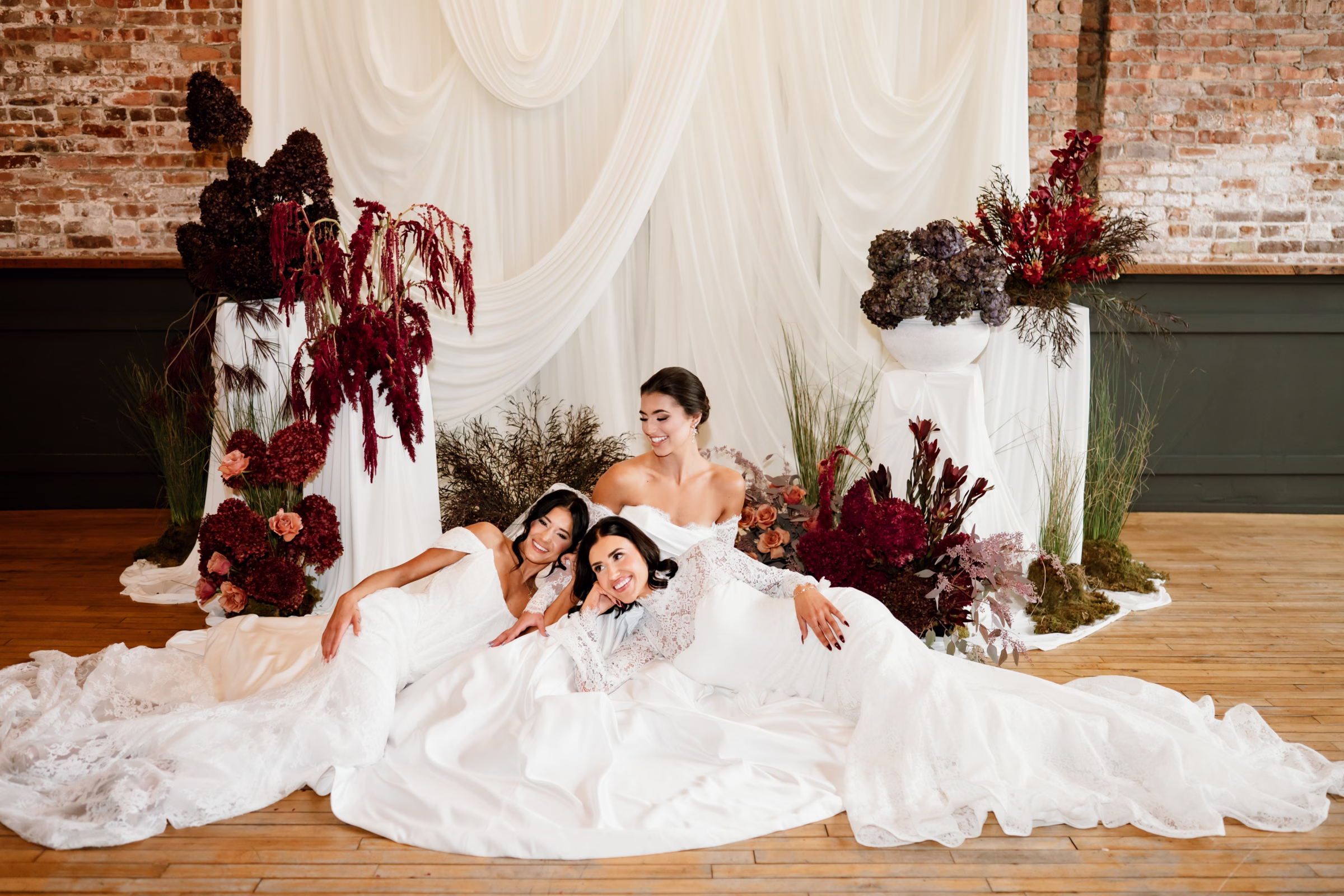 rich velvet fall wedding inspiration mercantile hall burlington wisconsin ceremony draping bride fashion 168 - Rich Velvet Fall Wedding Inspiration at Mercantile Hall in Burlington, Wisconsin three brides lounge in wedding dresses from Bon Bon Belle Bridal Boutique in front of a fall wedding ceremony backdrop at Mercantile Hall in Burlington, Wisconsin. The ceremony includes white drapery from Dream House Events LLC and burgundy and green florals from The Flower Bar and Anna's Florals