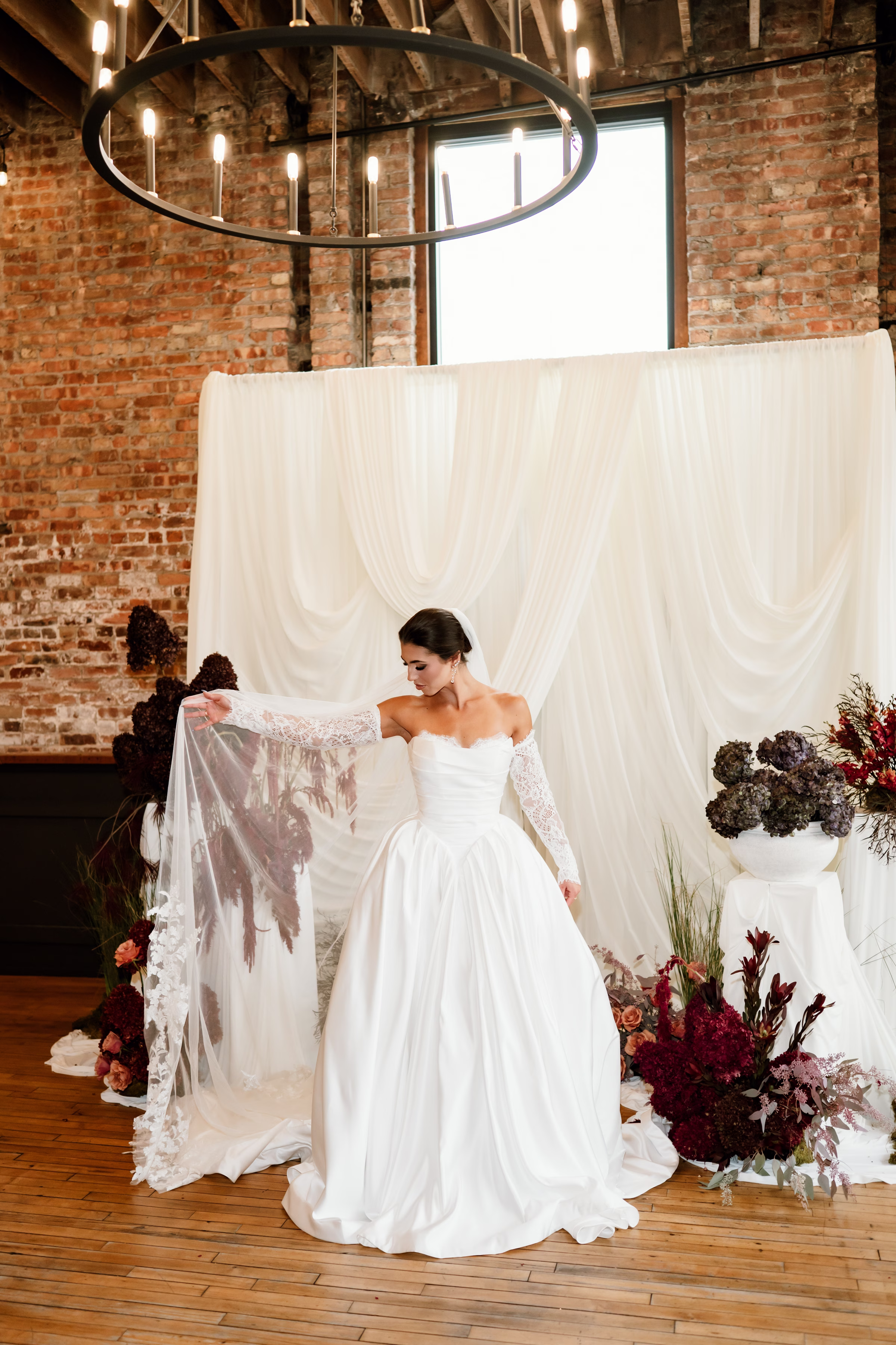 rich velvet fall wedding inspiration mercantile hall burlington wisconsin ceremony draping bride fashion 192 - Rich Velvet Fall Wedding Inspiration at Mercantile Hall in Burlington, Wisconsin a bride model poses at a wedding ceremony backdrop, showing off her dress and veil from Bon Bon Belle Bridal Boutique. Her strapless gown has a sweetheart neckline with peekaboo lace, a basque waist, princess skirt, and removable long lace sleeves. She is holding out a tulle veil with embroidered edges