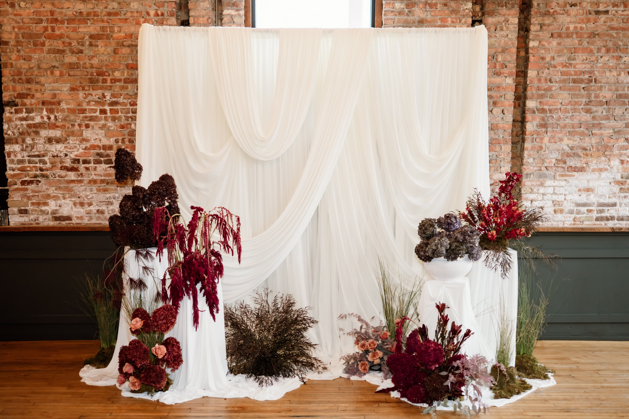 rich velvet fall wedding inspiration mercantile hall burlington wisconsin ceremony draping details 200 - Rich Velvet Fall Wedding Inspiration at Mercantile Hall in Burlington, Wisconsin a white curtain wedding ceremony backdrop by Dream House Events LLC surrounded by florals placed on the floor and on pillars. Prominent florals include hydrangeas, terracotta garden roses, cymbidium orchids, leucadendron, and ornamental grasses, accented with earthy moss and textural dried botanicals.