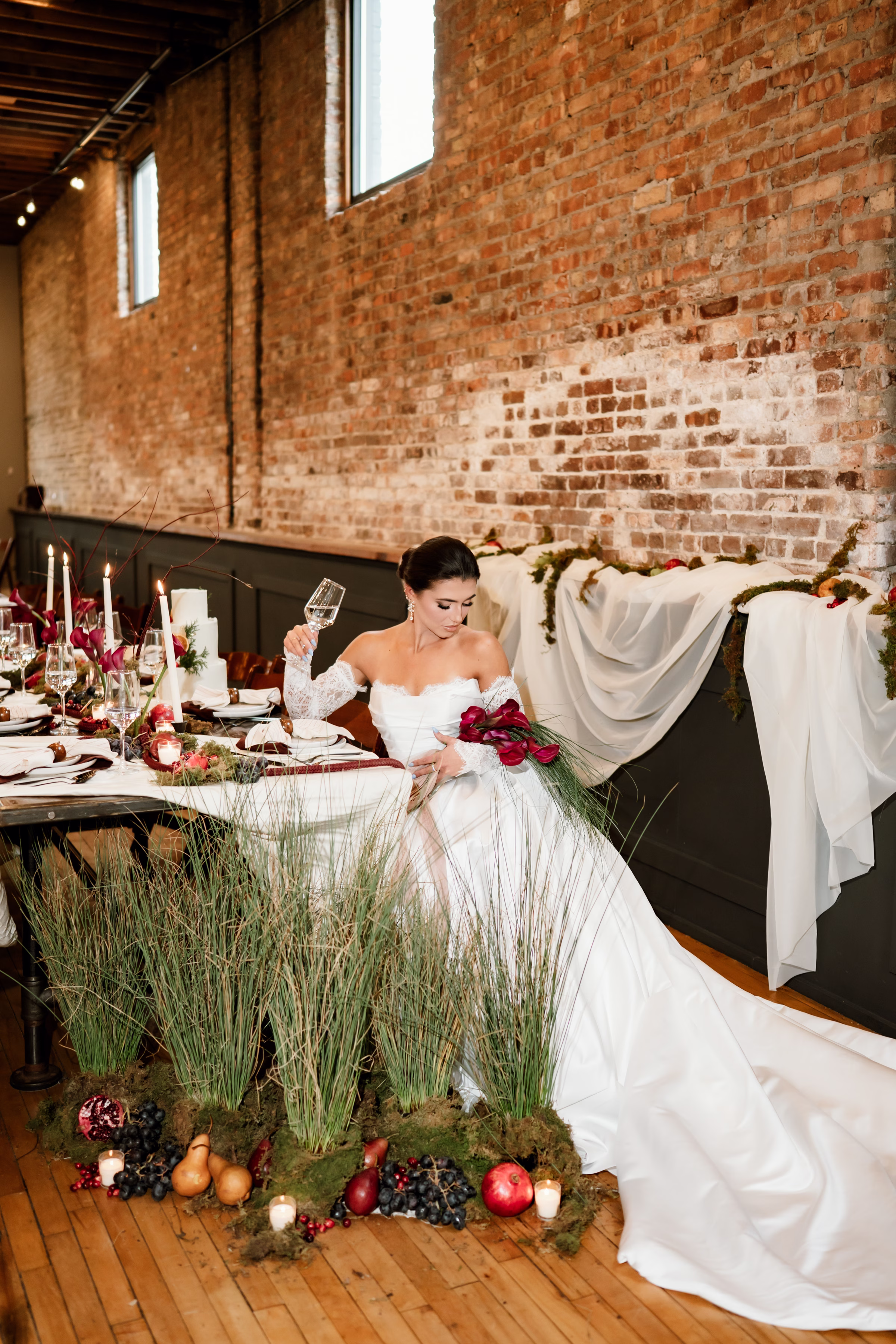rich velvet fall wedding inspiration mercantile hall burlington wisconsin tablescape bride florals 61 - Rich Velvet Fall Wedding Inspiration at Mercantile Hall in Burlington, Wisconsin a bride sits at a fall wedding tablescape, holding a wine glass in one hand and bouquet in the other. She is wearing a satin dress from Bon Bon Belle Bridal Boutique with a cathedral-length train