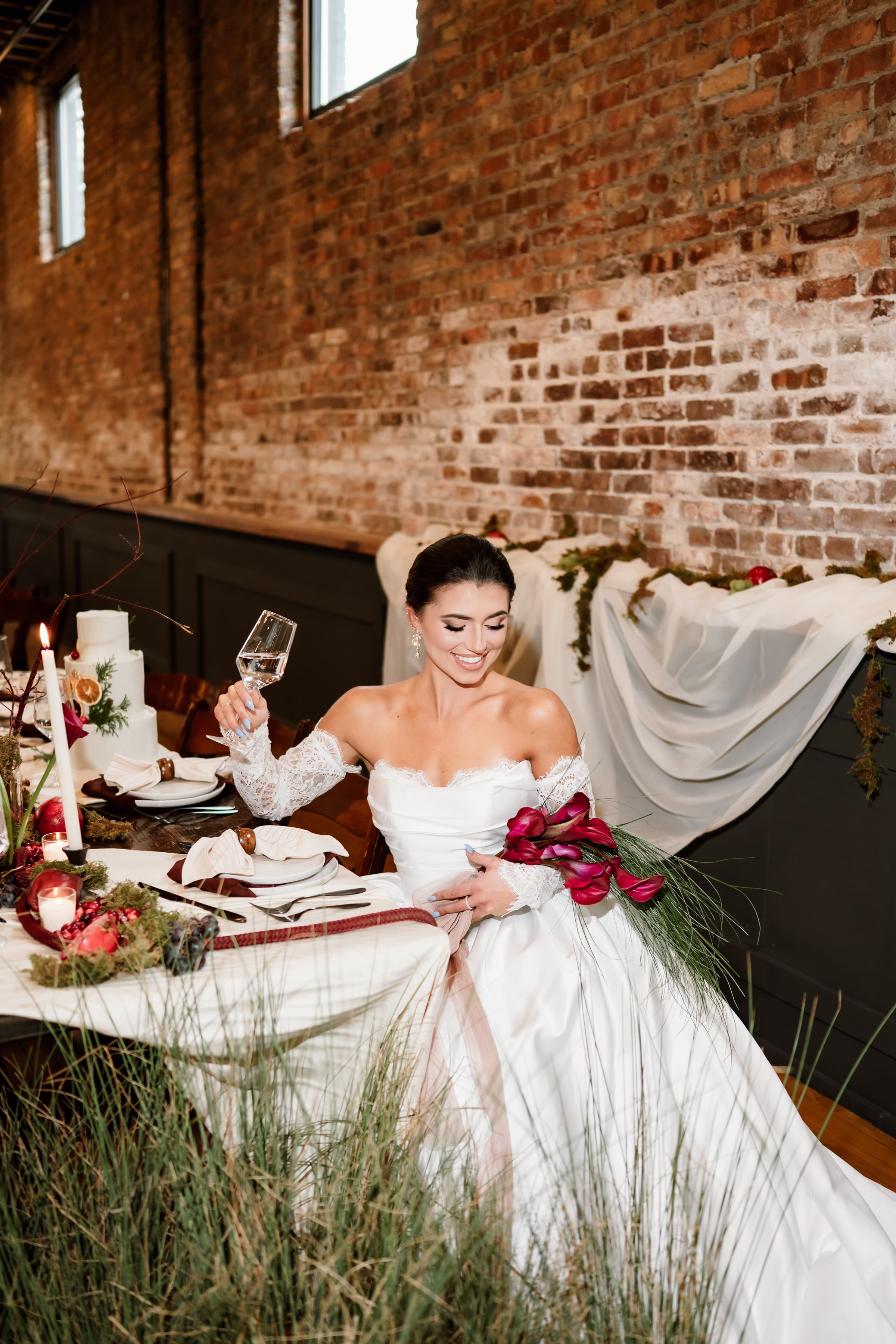rich velvet fall wedding inspiration mercantile hall burlington wisconsin tablescape bride florals 69 - Rich Velvet Fall Wedding Inspiration at Mercantile Hall in Burlington, Wisconsin a bride smiles, holding a wine glass in one hand and a bouquet of calla lilies with cascading bear grass. She is seated at a tablescape with a natural brick backdrop at Mercantile Hall wedding venue