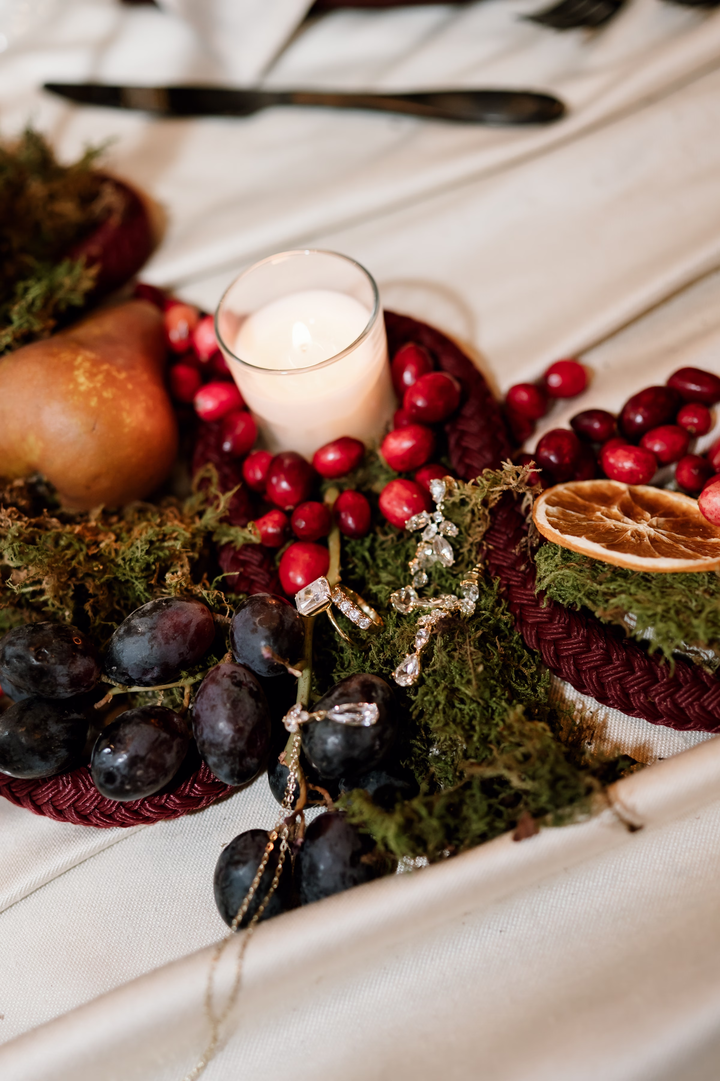 rich velvet fall wedding inspiration mercantile hall burlington wisconsin tablescape decor florals 41 - Rich Velvet Fall Wedding Inspiration at Mercantile Hall in Burlington, Wisconsin a close-up of a fall wedding tablescape centerpiece showing jewelry placed on mossy greenery surrounded by a glowing candle, purple grapes, cranberries, a pear, and an orange slice